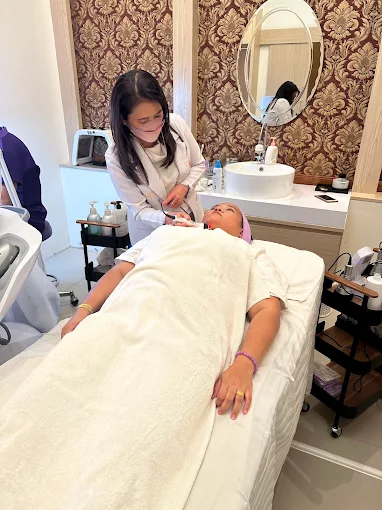 พนักงานทำทรีตเมนต์ผิวหน้าให้ลูกค้าที่นอนห่มผ้าบนเตียง โดยมีอ่างล้างหน้าและกระจกอยู่ด้านหลัง